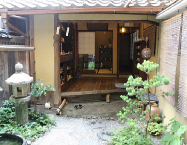 The entrance of a Kyoto machiya: a gravel garden with a stone lantern and a small pine, and beyond it the raised wooden floor, with a pair of sandals left on the stone step below it.