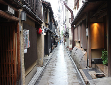 A narrow roji lane running back between two machiya, wooden lattice on both sides and the tiled roofline closing overhead.