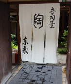 A dyed noren curtain hanging across the doorway of a Kyoto machiya, with potted plants on the stone step beneath it and the wooden lattice of the front visible either side.